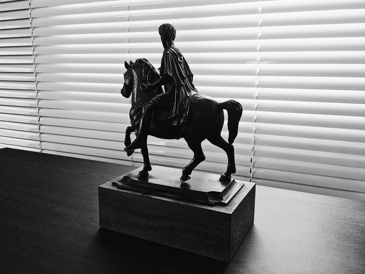 Black and white photo of a horse sculpture in front of window blinds.